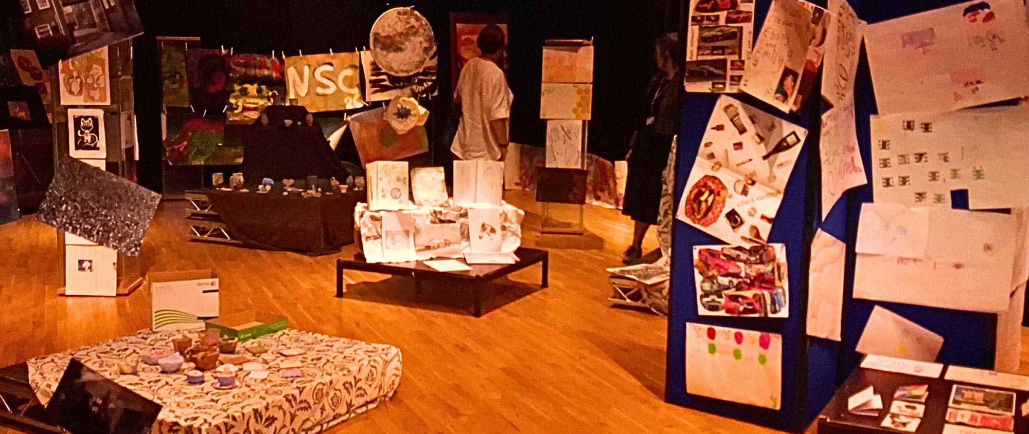 Exhibiliton space showing artwork in various media, created by young people, on walls and tables; two viewers are admiring the show.
